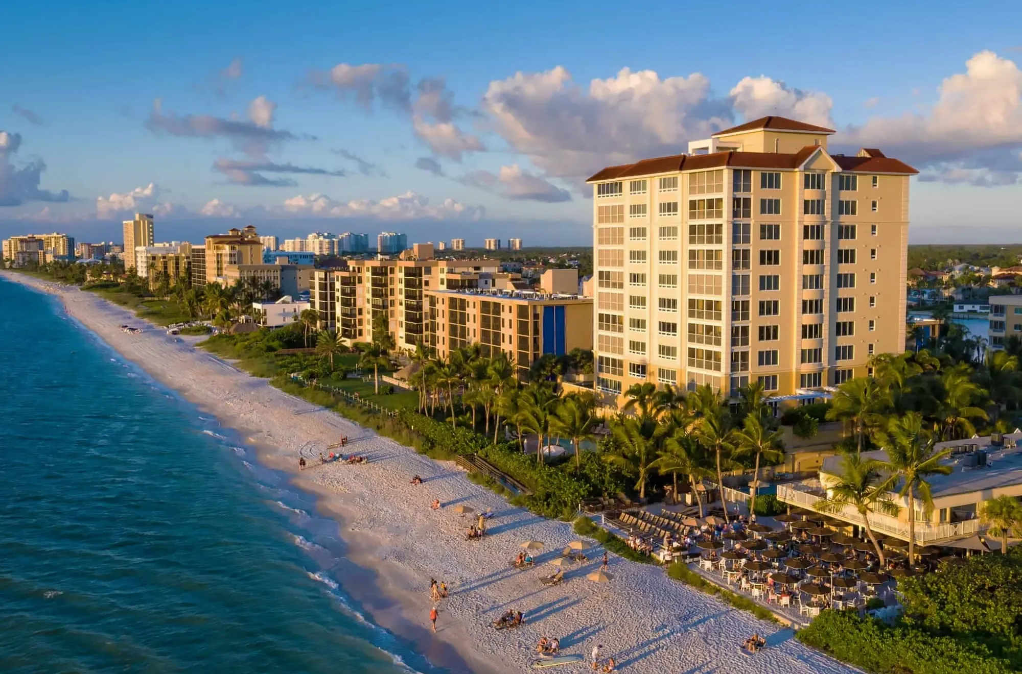 Aerial view of the Naples, Florida shoreline at golden hour with mid-rise condominium buildings, royal palms and beachgoers on white sand