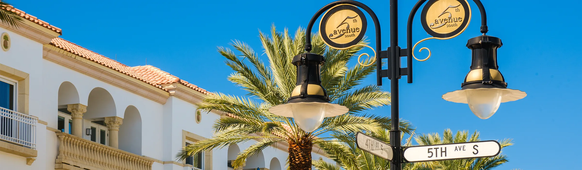 Fifth Avenue South street sign and lamppost at Fourth Street South, beside a Mediterranean-style building with arched balconies in downtown Naples, Florida