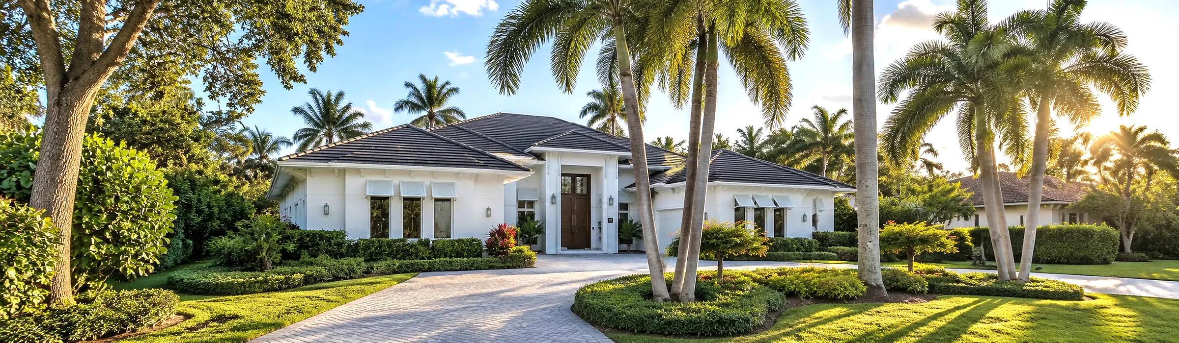 Single-story white luxury home with a dark tile roof, paver driveway and royal palms in a Naples, Florida neighborhood